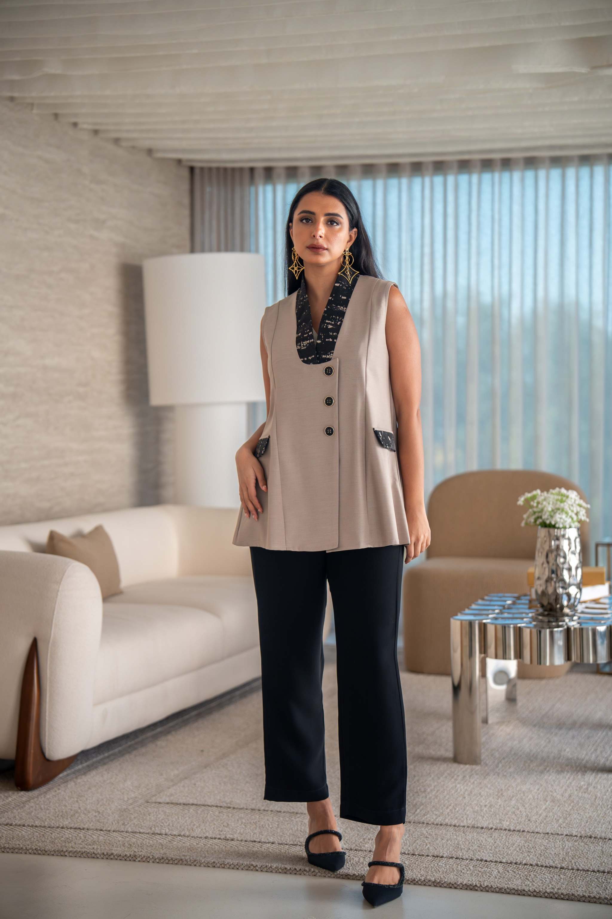Woman wearing beige vest with black printed lapel and decorative buttons, styled with black pants in modern living room.