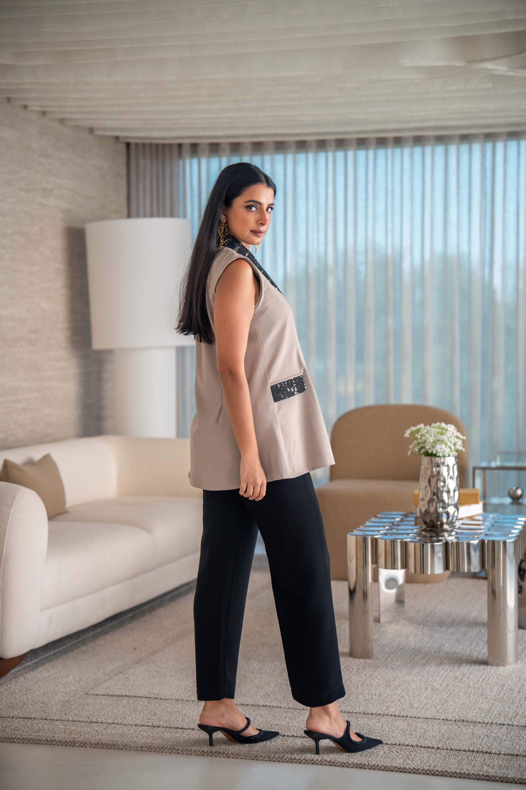 Beige vest with black printed lapel, decorative buttons, and angled side pockets, styled with black pants in a modern living room.