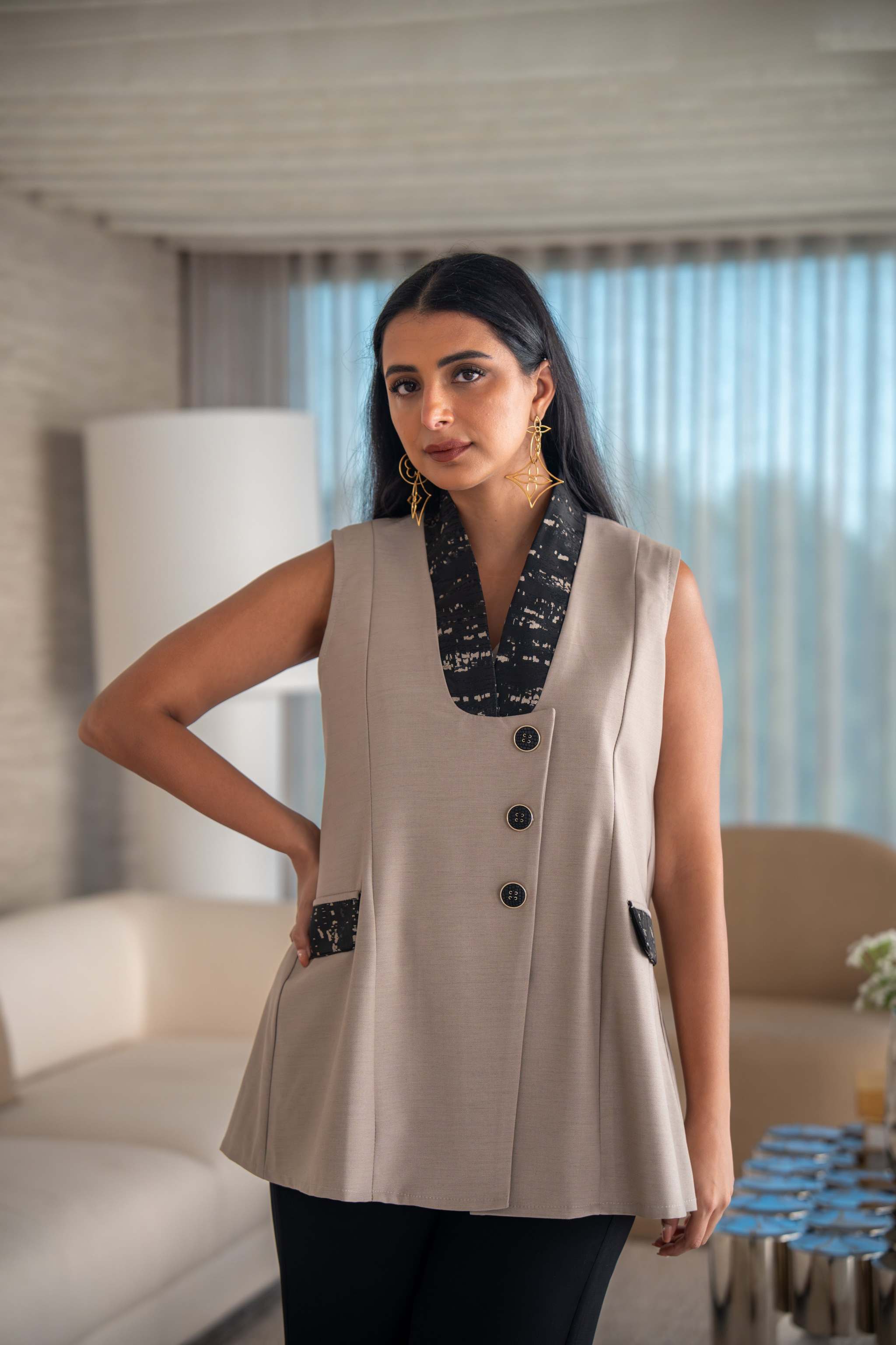 Beige vest with black printed lapel, decorative buttons, and angled side pockets, styled with black pants in a modern living room.