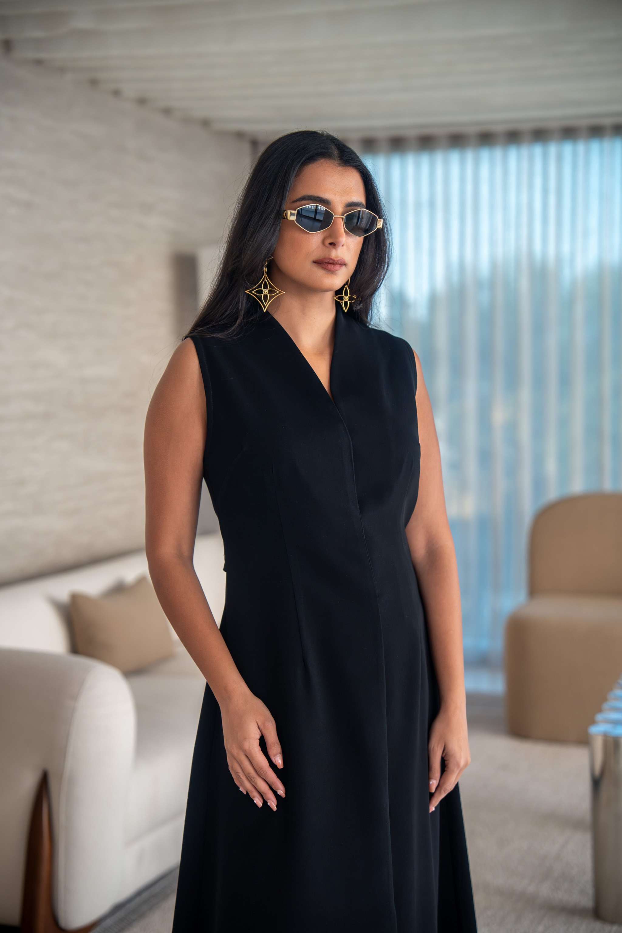 Woman wearing elegant black sleeveless crepe dress with v-neck and sunglasses in modern living room