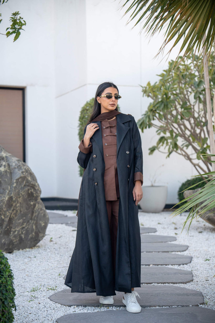Woman wearing espresso linen abaya with black longline trench coat-style blazer and white sneakers outdoors