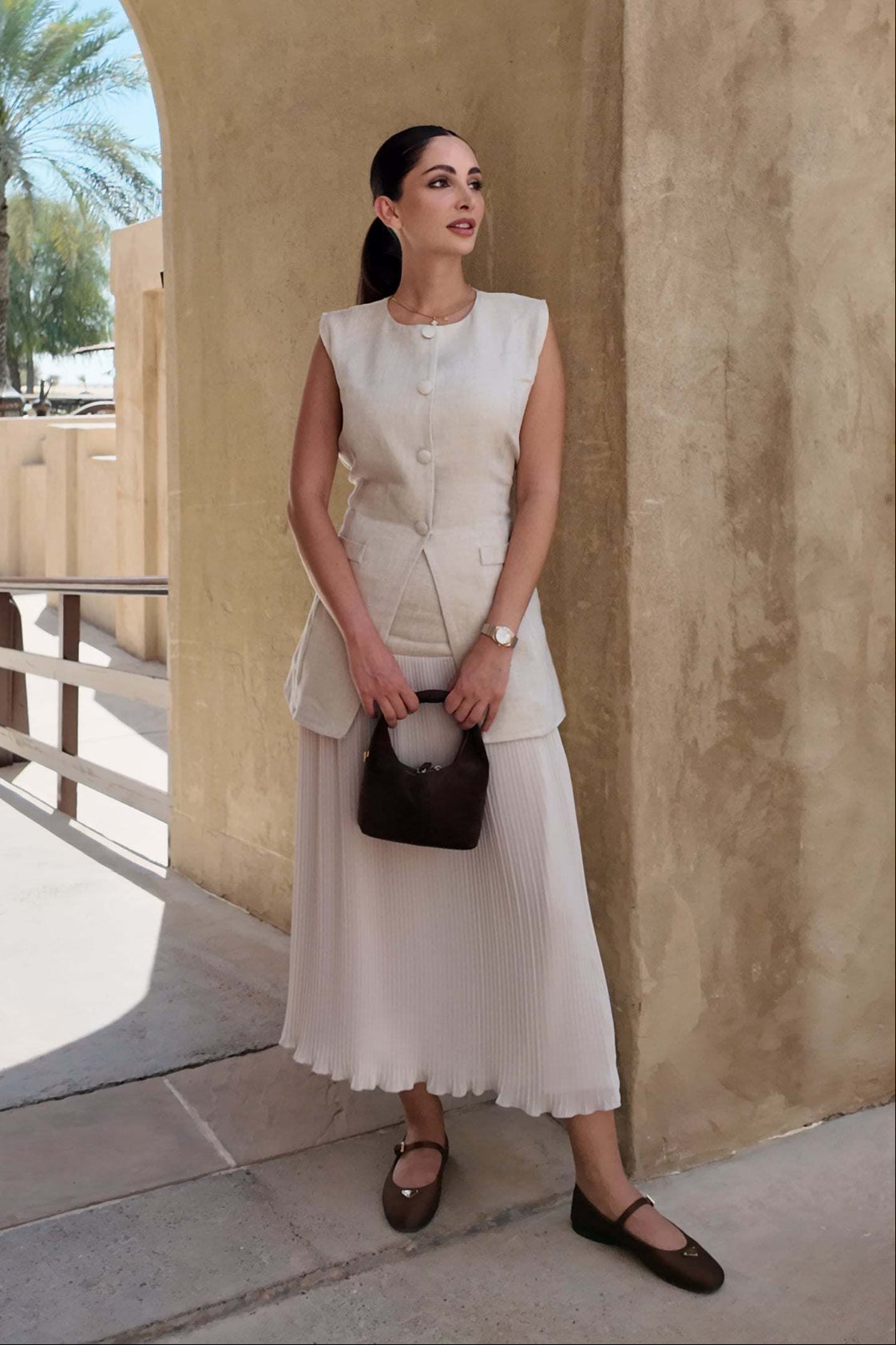 Sleeveless beige Sandy Pleats dress with tailored bodice, front buttons, cinched waist, and soft pleated skirt perfect for warm-weather occasions.