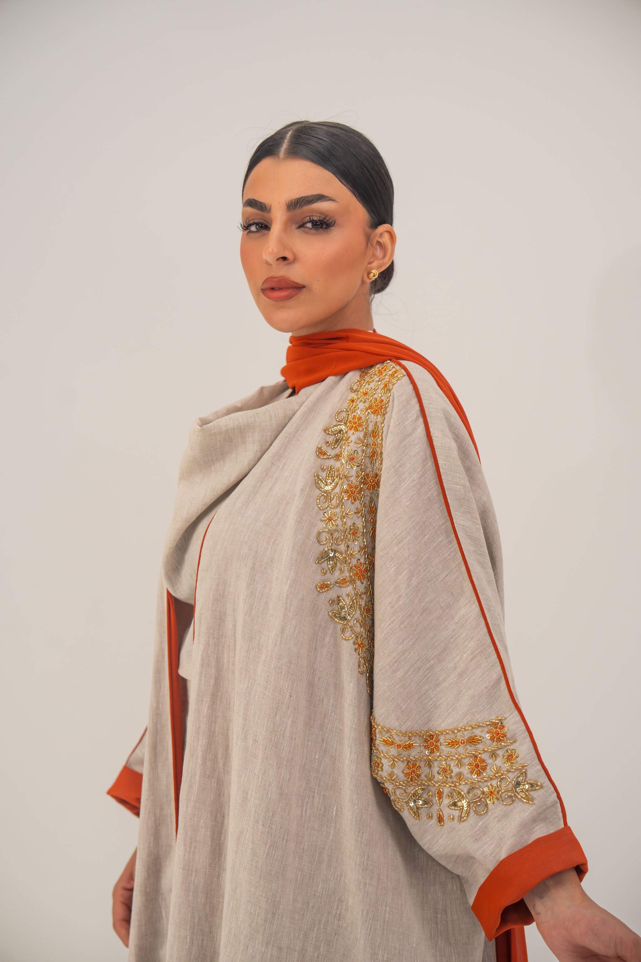 Woman wearing a beige linen-blend abaya with gold and orange beaded embellishments and orange chiffon lining on sleeves and collar