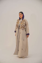 Woman wearing a beige open-front bisht abaya with gold and purple floral beadwork, purple chiffon lining, and relaxed fit.