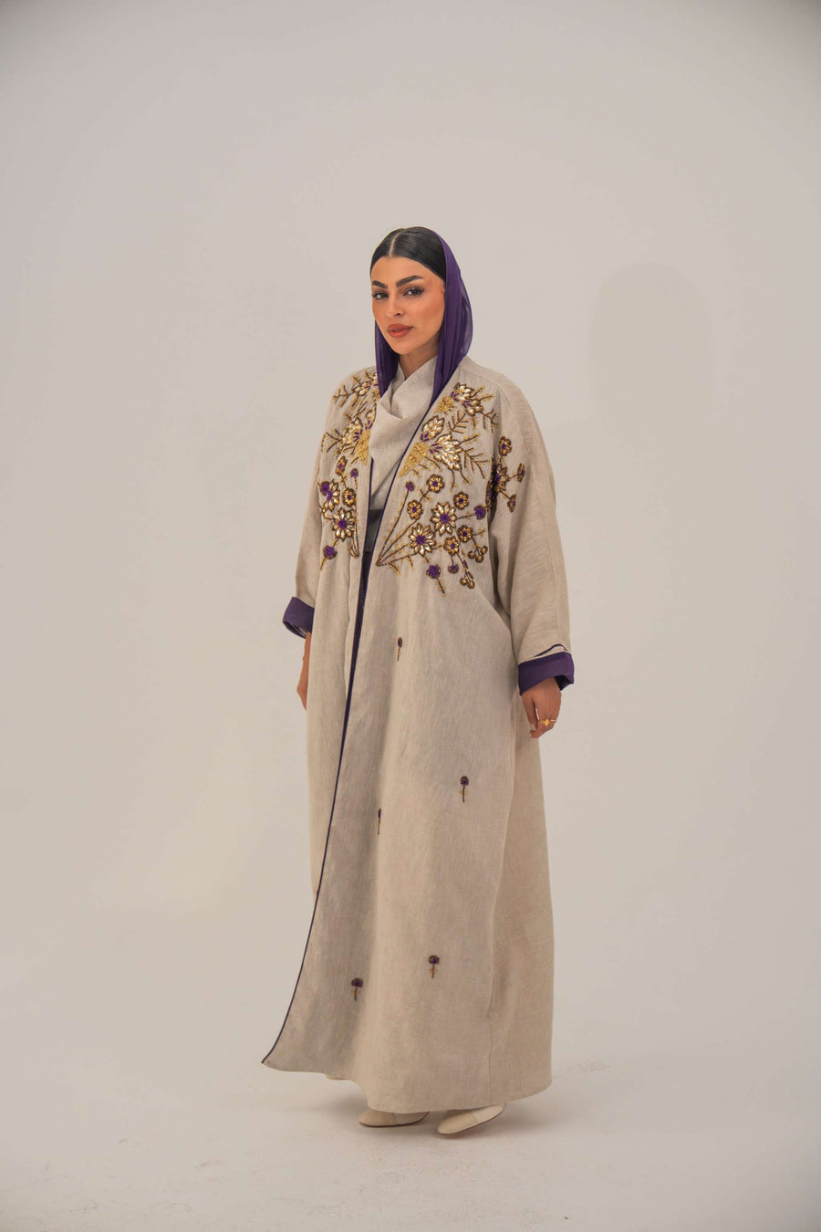 Woman wearing a beige open-front bisht abaya with gold and purple floral beadwork, purple chiffon lining, and relaxed fit.