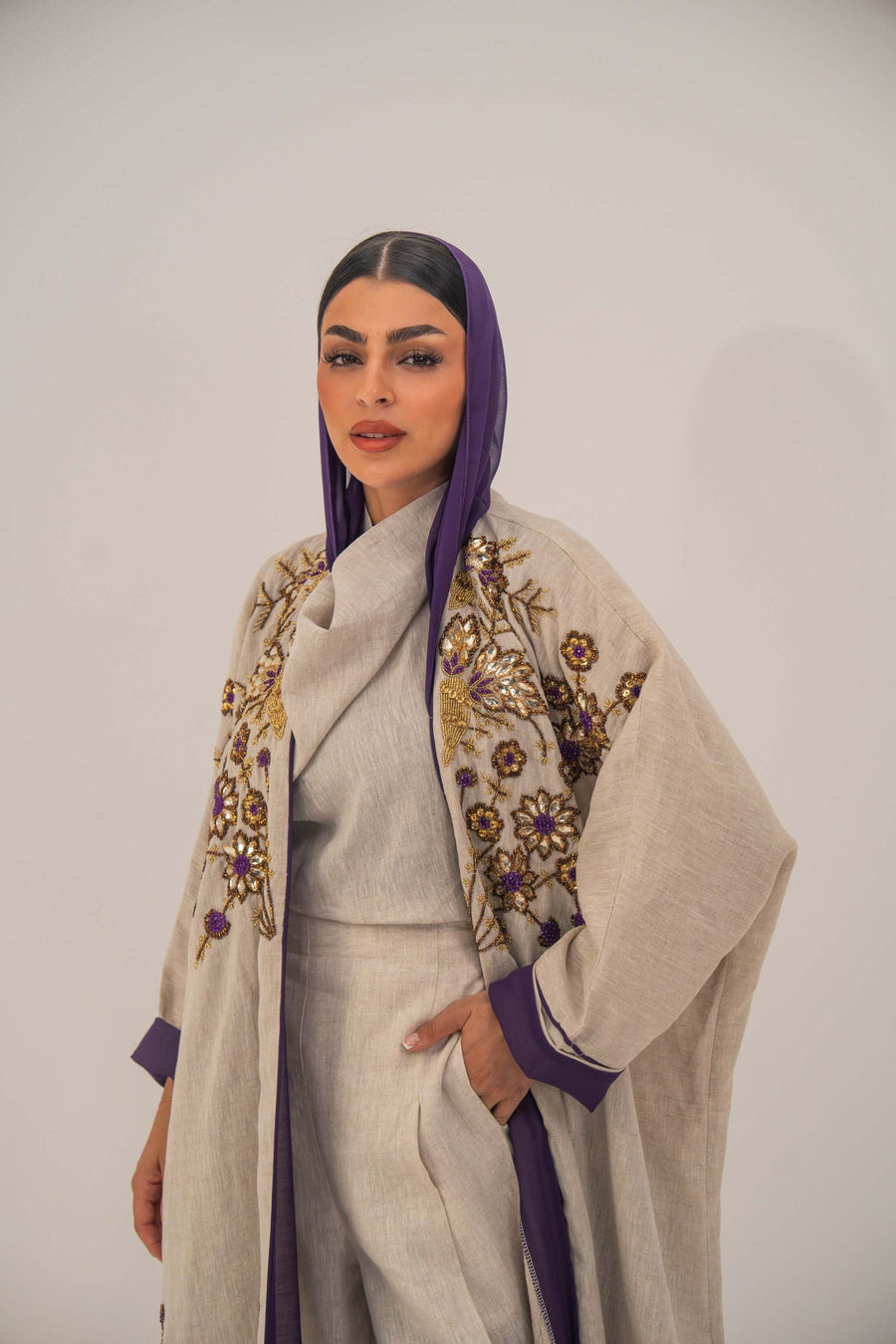 Woman wearing a beige open-front bisht abaya with gold and purple floral beadwork, purple chiffon lining, and relaxed fit.