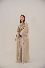 Woman wearing a beige open-front bisht abaya with gold and purple floral beadwork, purple chiffon lining, and relaxed fit.