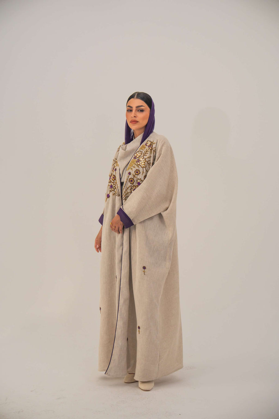 Woman wearing a beige open-front bisht abaya with gold and purple floral beadwork, purple chiffon lining, and relaxed fit.