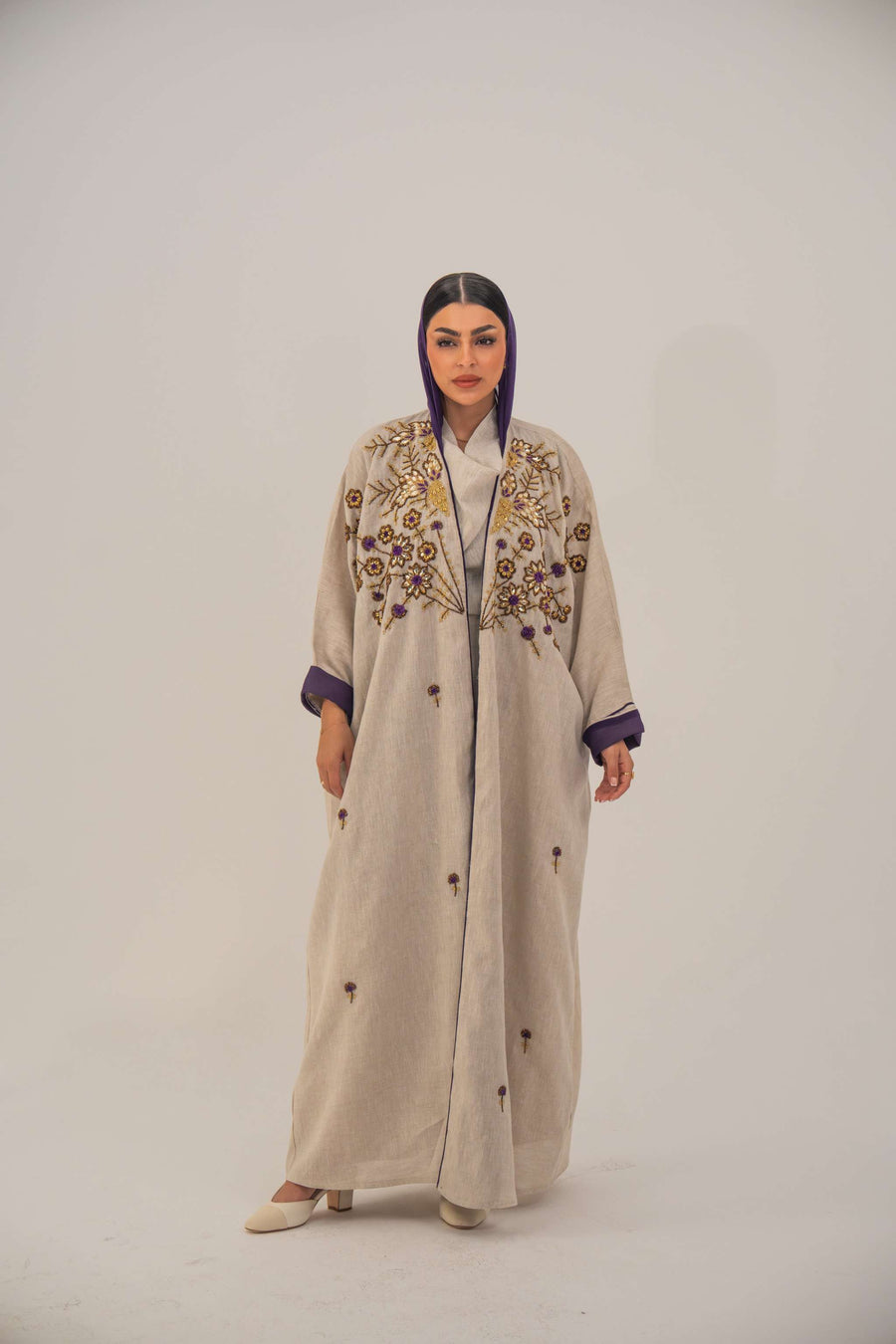 Woman wearing a beige open-front bisht abaya with gold and purple floral beadwork, purple chiffon lining, and relaxed fit.