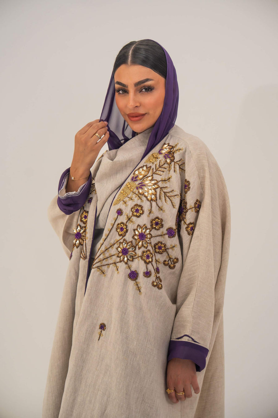 Woman wearing a beige open-front bisht abaya with gold and purple floral beadwork, purple chiffon lining, and relaxed fit.