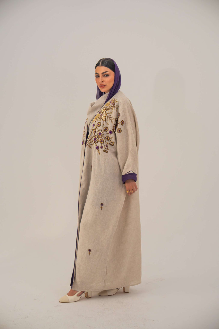 Woman wearing a beige open-front bisht abaya with gold and purple floral beadwork, purple chiffon lining, and relaxed fit.