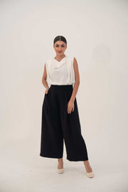 Woman wearing a sleeveless white blouse and wide black pants standing against a plain light background