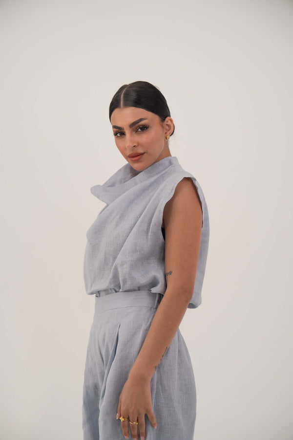 Woman wearing sleeveless light blue linen blouse with draped collar and relaxed fit.