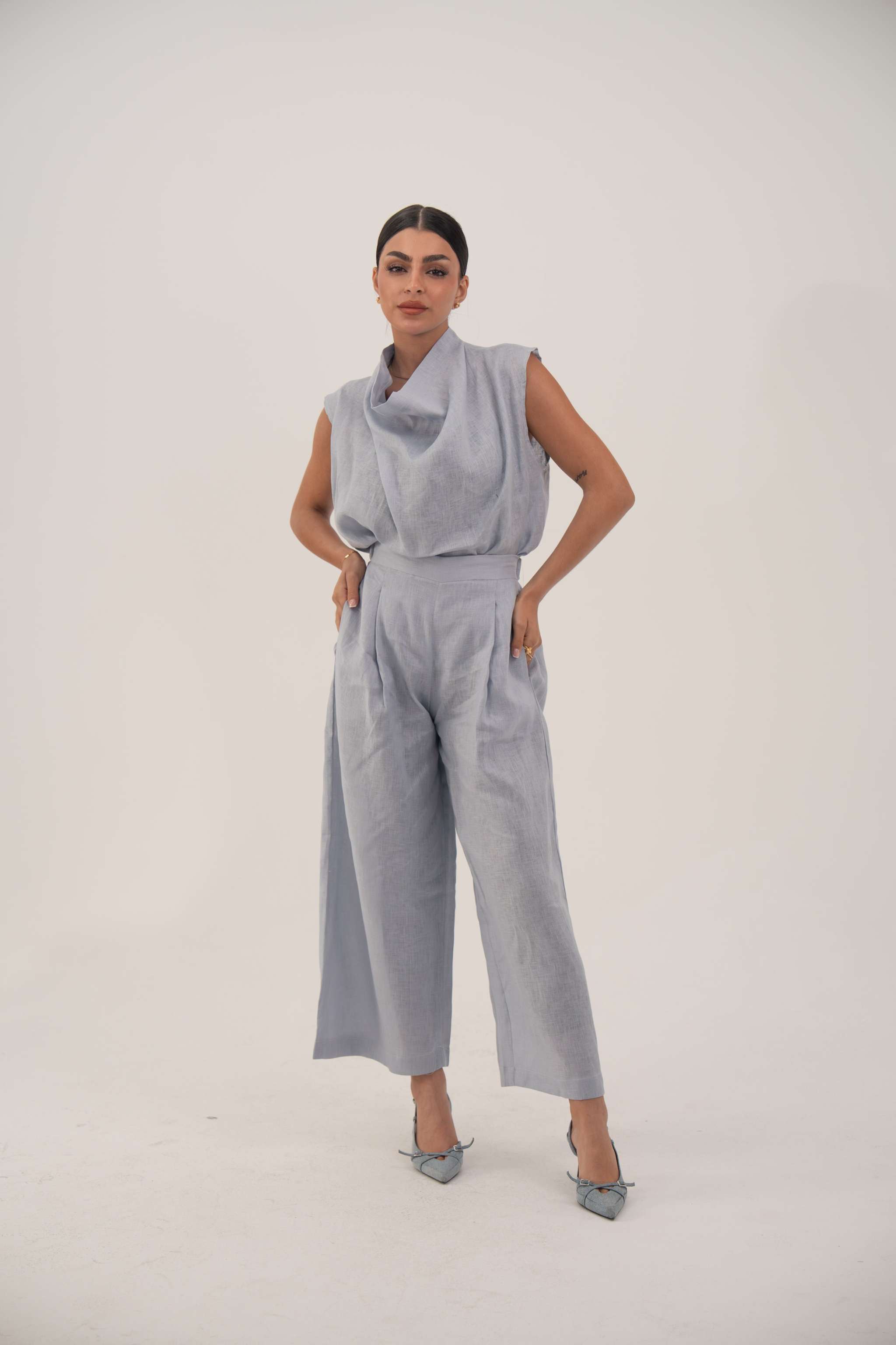 Woman wearing a sleeveless light blue linen blouse with a draped collar and relaxed fit paired with matching wide-leg pants.