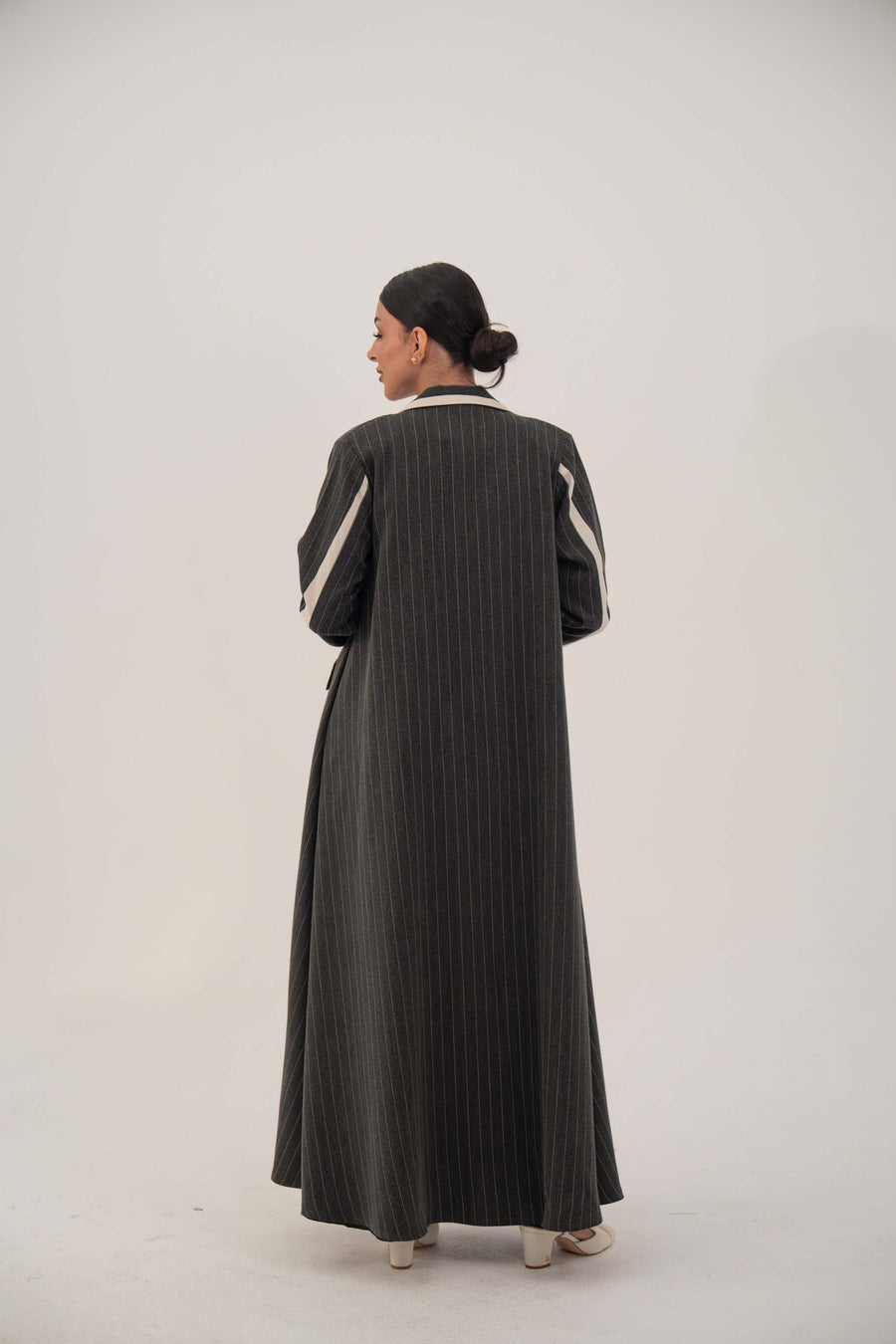 Back view of a woman wearing a dark pinstriped blazer-style abaya with off-white inner cuffs and shoulder details, standing against a plain background.