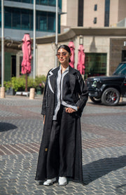 Woman wearing black blazer abaya with sharp lapels and white piping, styled for modern street and professional look.