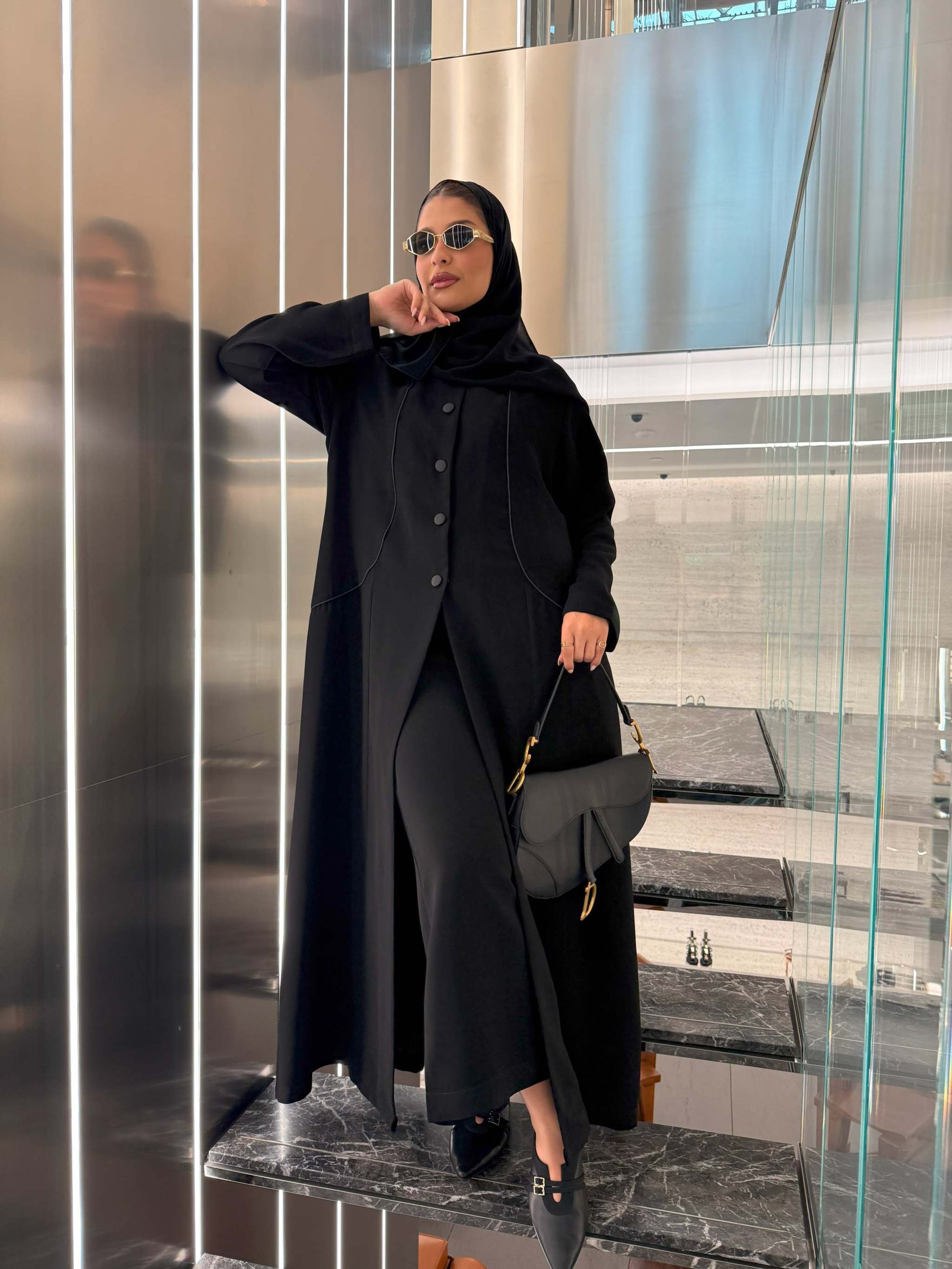 Black crepe blazer-style abaya with silk lining, structured buttons, and subtle seam detailing worn with sunglasses and black handbag.