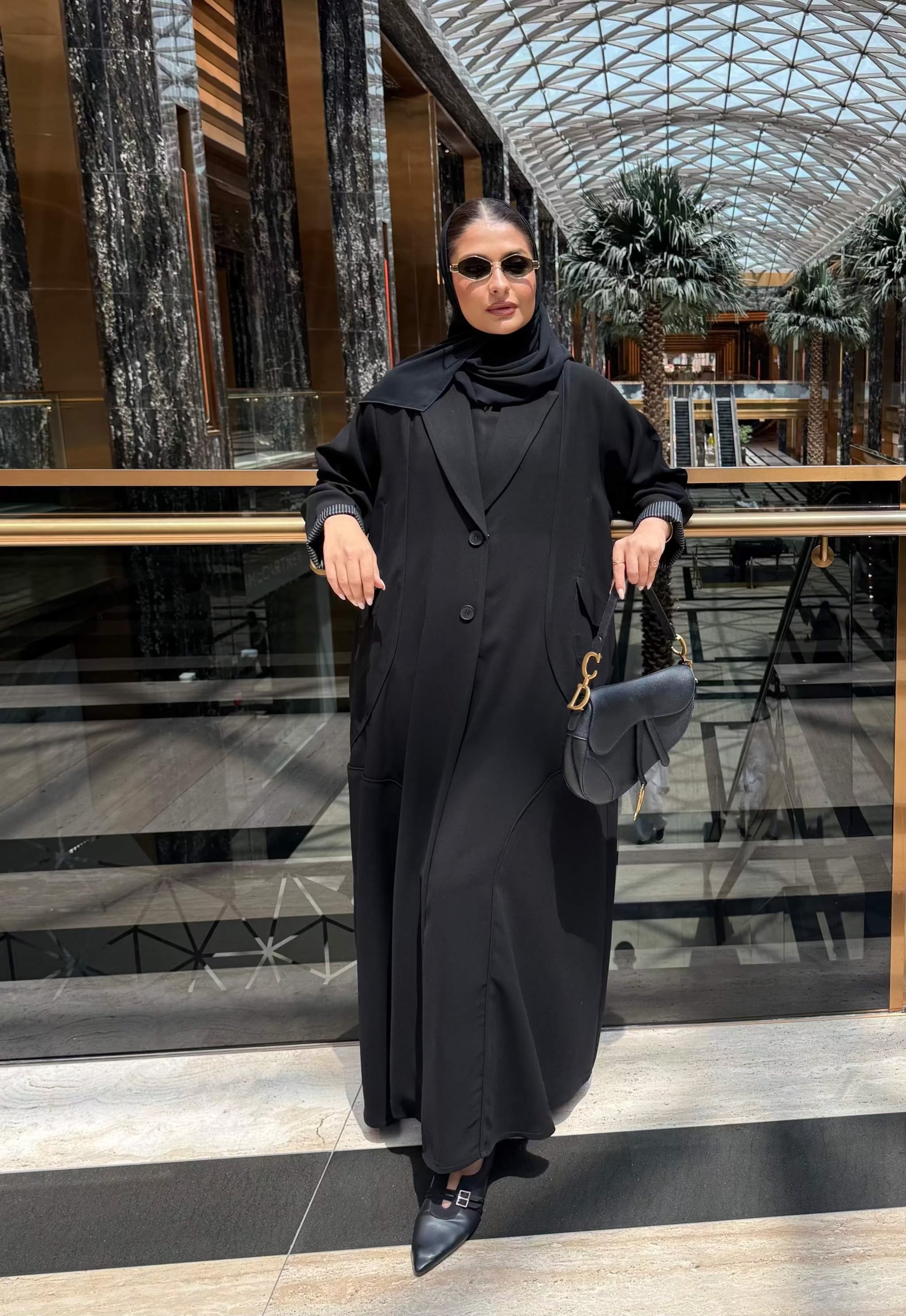 Black button-down crepe abaya with relaxed fit and side pockets worn by woman with sunglasses and handbag