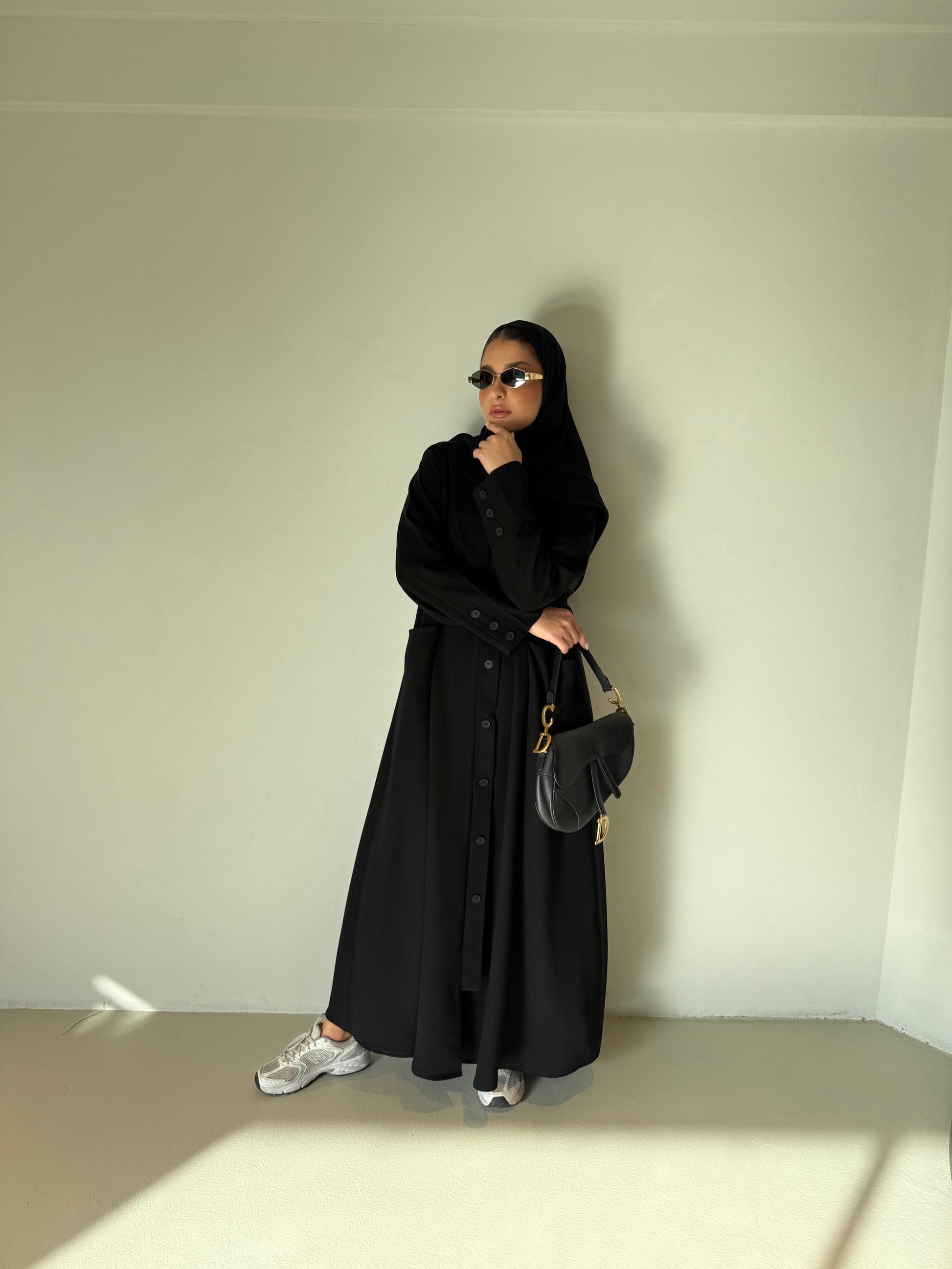 Woman wearing The Flowing Crepe Button-Down Abaya in black with sunglasses and handbag standing against a plain wall
