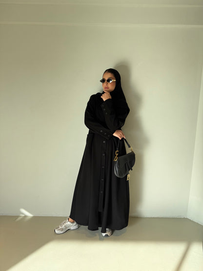 Woman wearing The Flowing Crepe Button-Down Abaya in black with sunglasses and handbag standing against a plain wall
