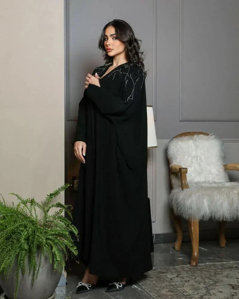 Woman wearing black crepe bisht with silver and black hand embroidery on front and sleeves standing indoors next to a lamp and plant.