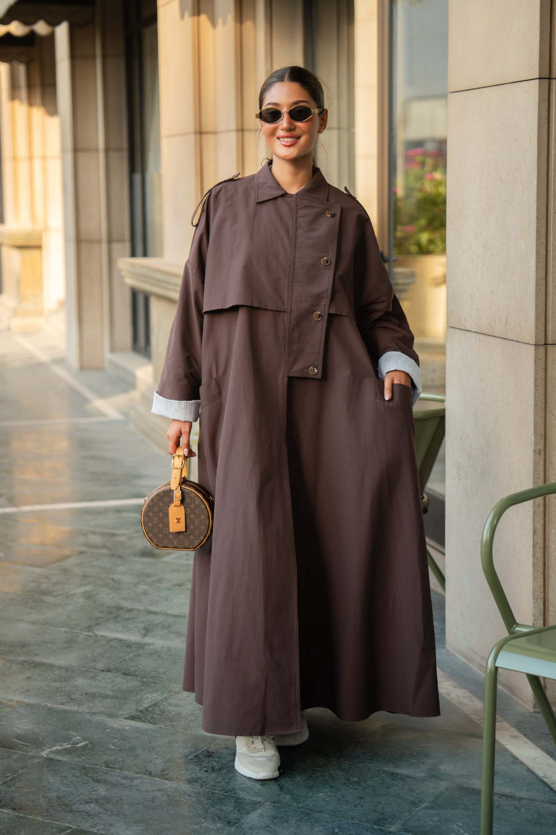 Woman wearing chocolate brown trench coat abaya with button-down front and layered detailing, styled with sunglasses and white sneakers outdoors
