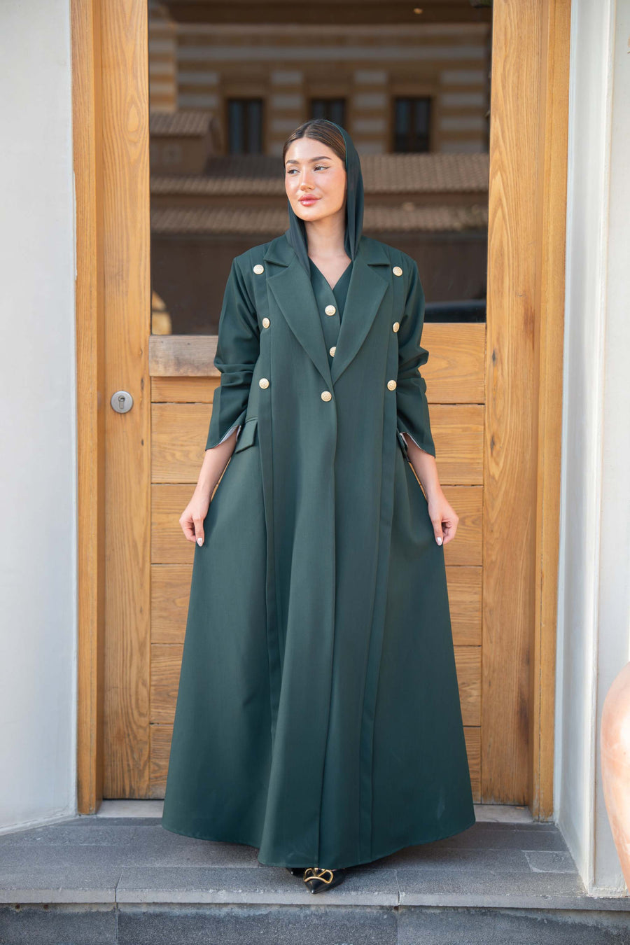 woman wearing emerald green double-breasted abaya with gold-tone buttons and structured lapel collar standing in front of wooden door