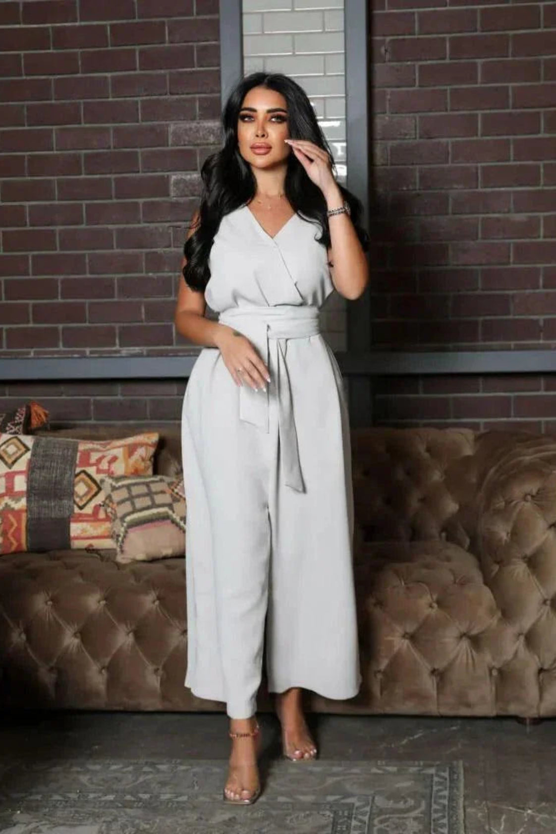 woman wearing sleeveless grey jumpsuit with wrap V-neckline and waist tie standing in front of brick wall