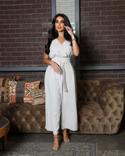 woman wearing sleeveless grey jumpsuit with wrap V-neckline and waist tie standing in front of brick wall
