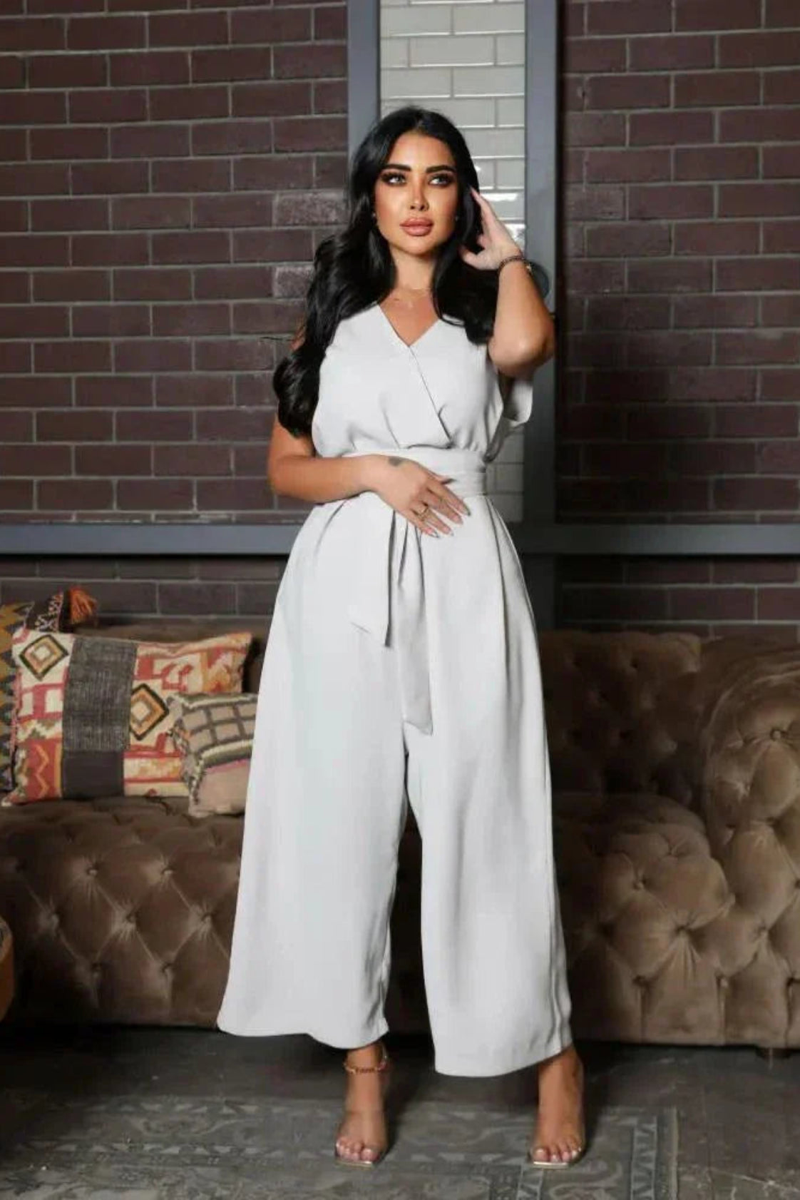 woman wearing sleeveless grey jumpsuit with wrap V-neckline and waist tie standing in front of brick wall