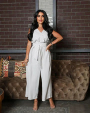 woman wearing sleeveless grey jumpsuit with wrap V-neckline and waist tie standing in front of brick wall