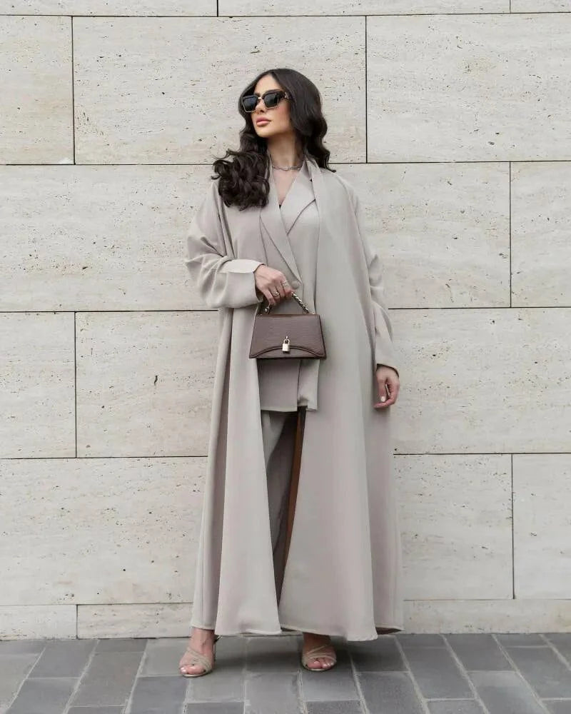 Woman wearing a flowing soft beige crepe outfit with lightweight textured fabric, holding a handbag and sunglasses, standing against a stone wall.