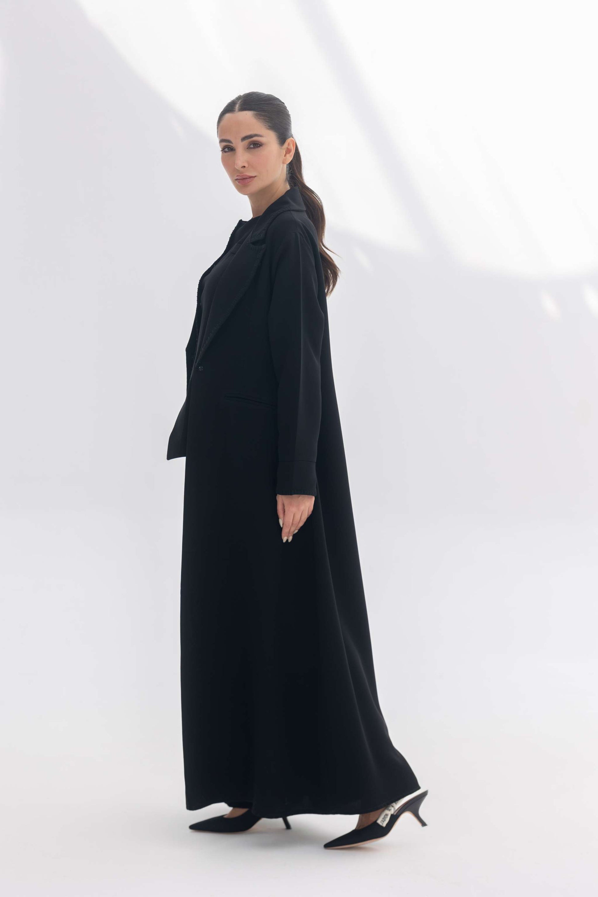 Woman wearing a black tailored crepe blazer abaya with intricate embroidery and matching heels in a minimalistic setting