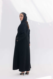 Woman wearing a sophisticated black crepe blazer abaya with tailored silhouette and black embroidery, paired with a matching black hijab and heels.