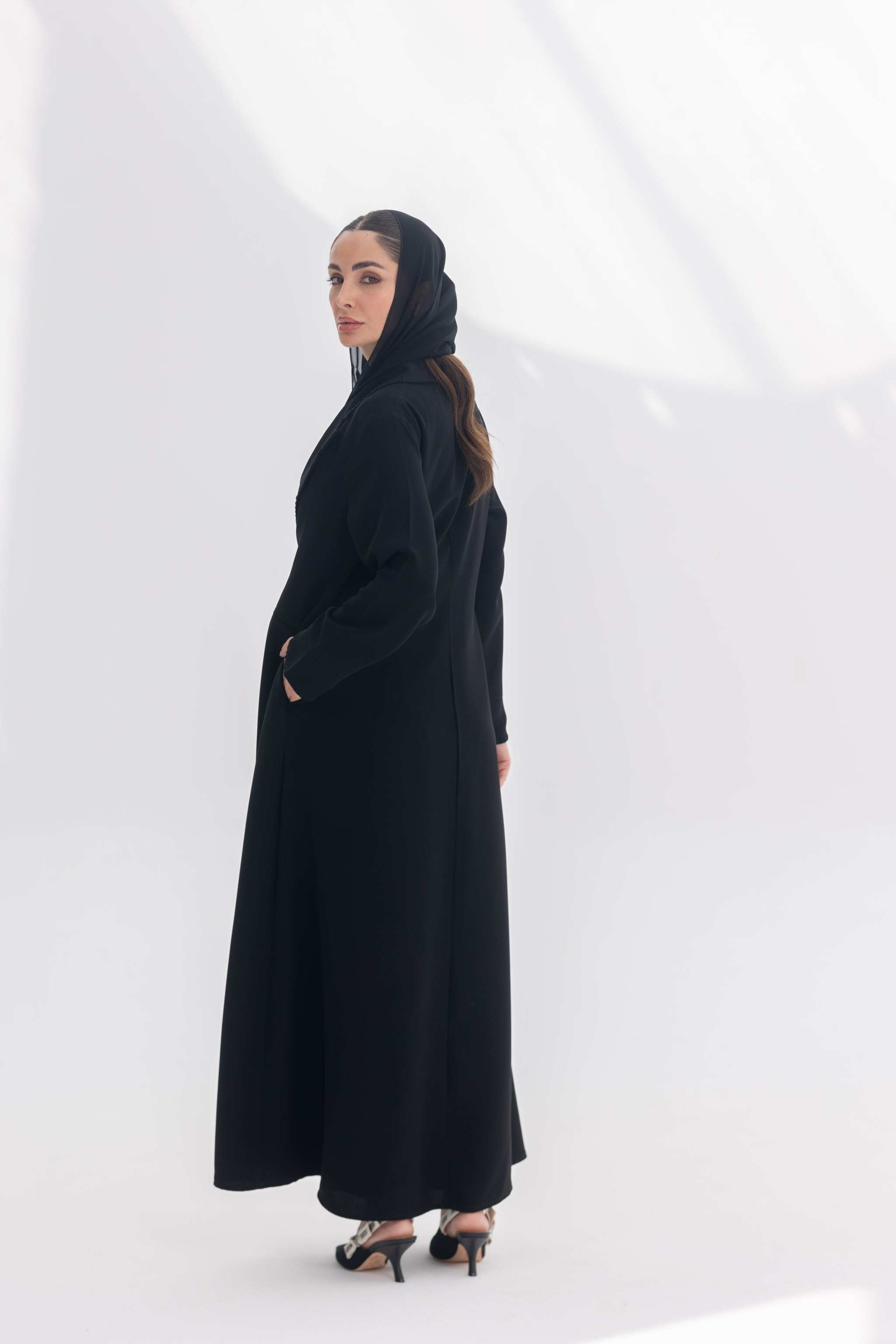 Woman wearing a sophisticated black crepe blazer abaya with tailored silhouette and black embroidery, paired with a matching black hijab and heels.