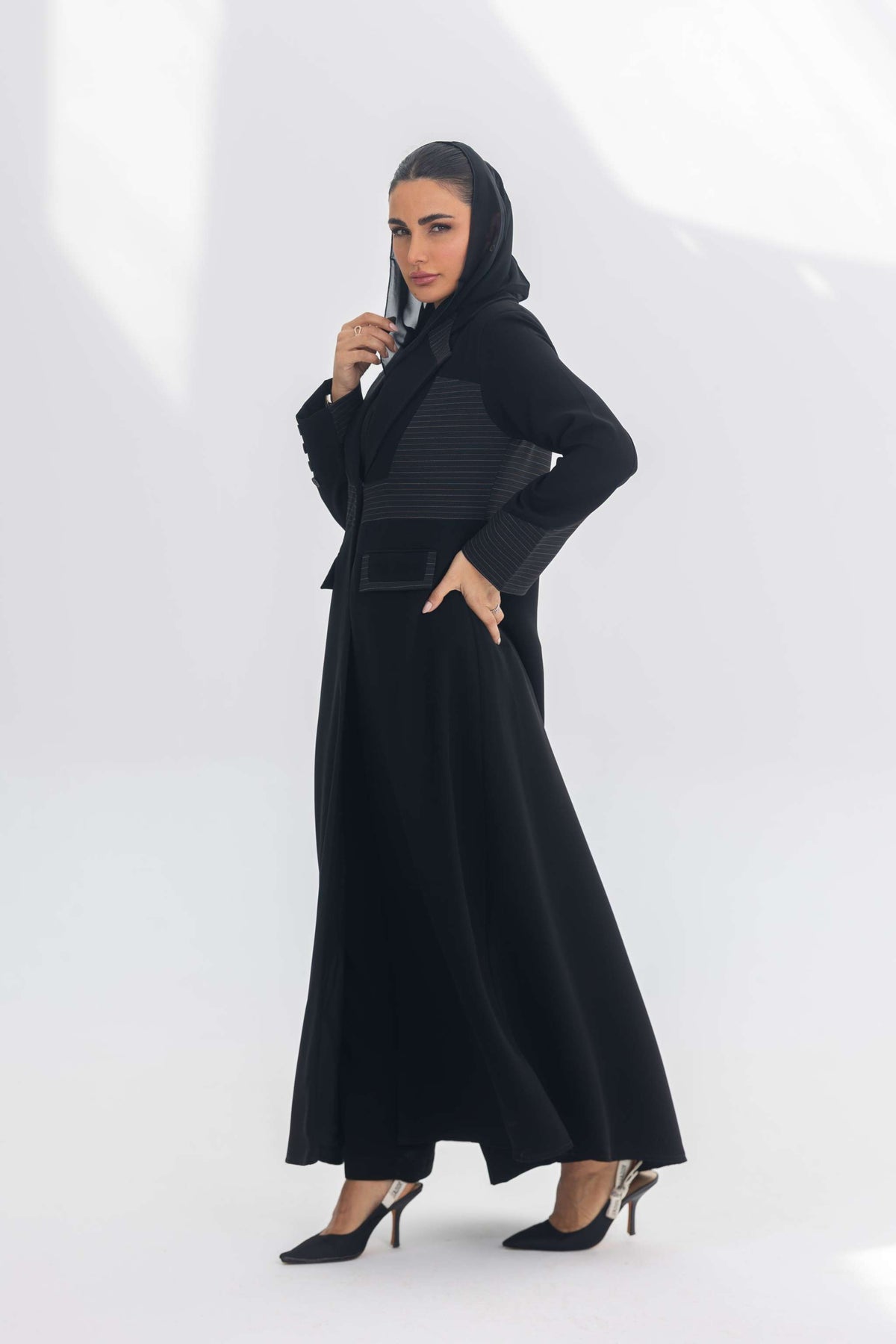 Woman wearing black crepe blazer abaya with contrasting fabric detailing and hood, standing in elegant pose against white background