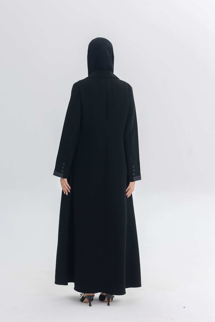 Back view of a woman wearing a black Denim Veil abaya with a hood and long sleeves in a minimal studio setting