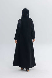 Back view of a woman wearing a black satin and crepe abaya with lace details and a hood, standing against a plain white background.