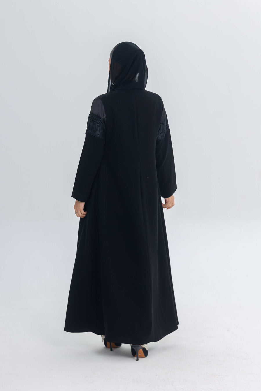 Back view of a woman wearing a black satin and crepe abaya with lace details and a hood, standing against a plain white background.