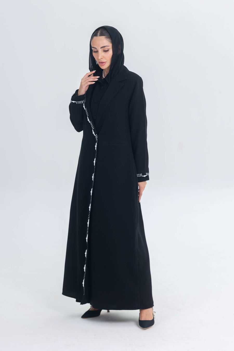 Black crepe blazer abaya with white pearl and bead embroidery, tailored silhouette, worn by a woman with a headscarf and black heels.