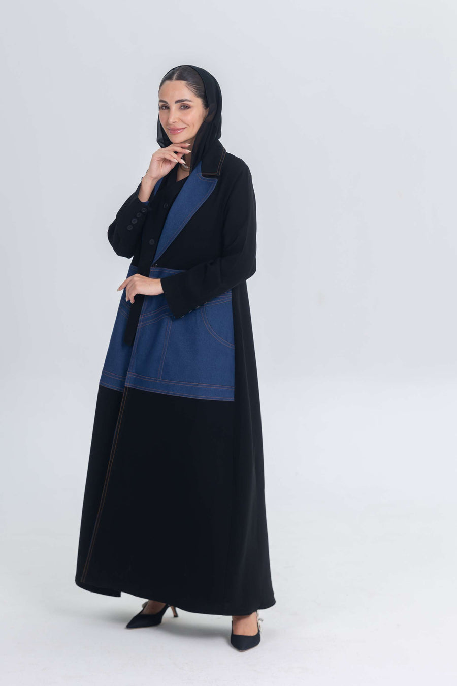 Woman wearing black crepe abaya with denim panels and black hijab standing against white background