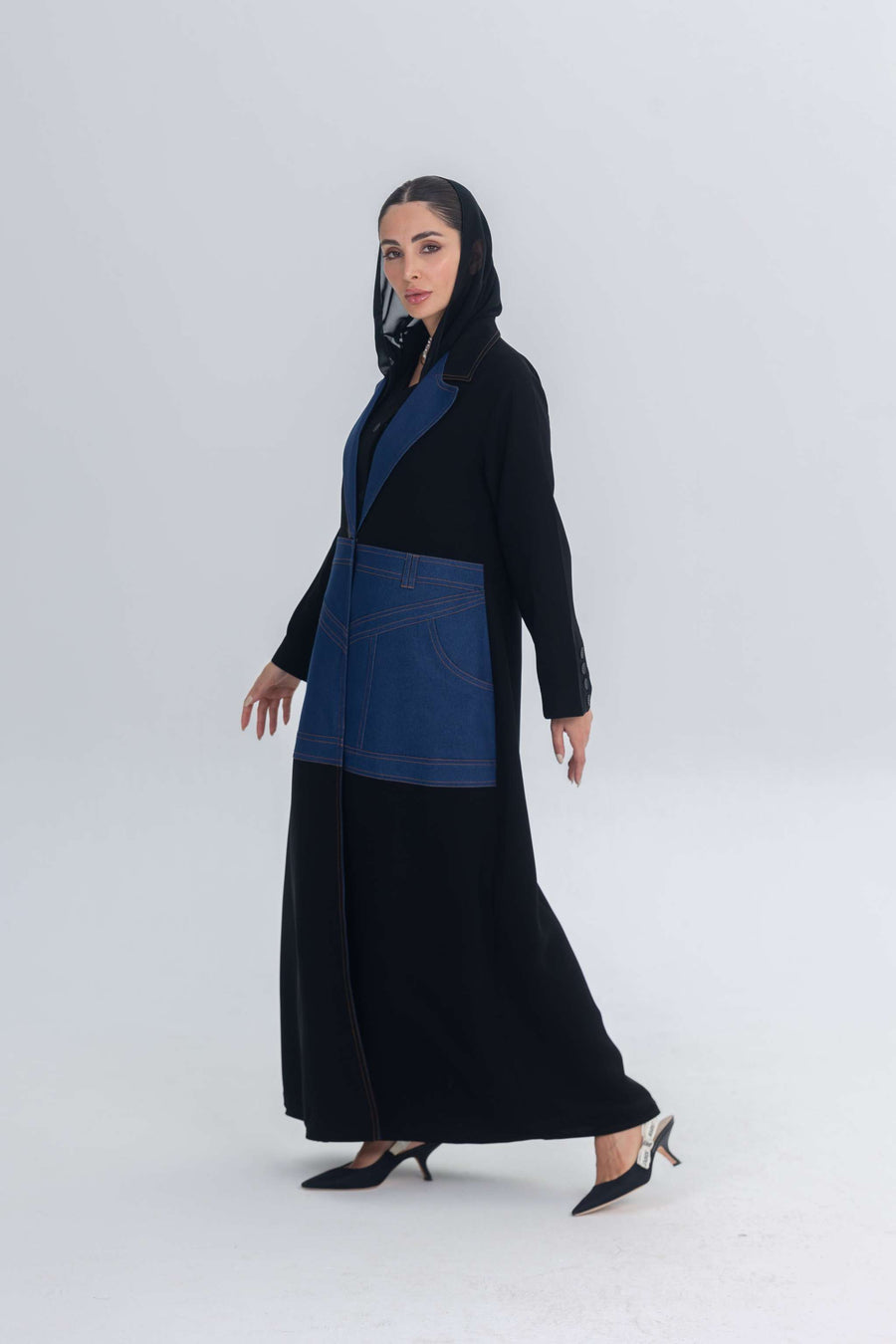 Black crepe abaya with blue denim panels and hood worn by woman in heels against light background