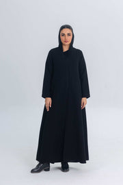 Woman wearing black crepe abaya with subtle satin detailing and hood, standing against plain background