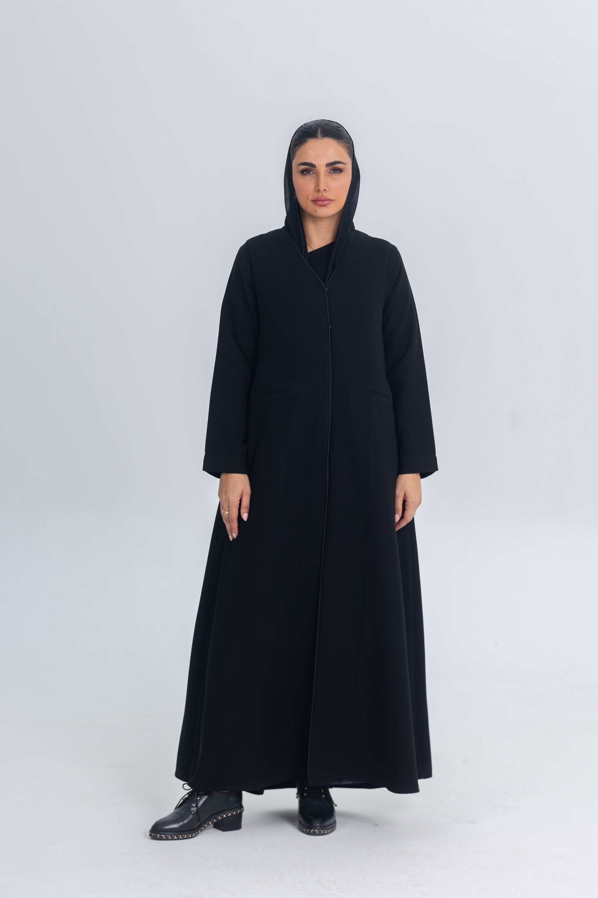 Woman wearing black crepe abaya with subtle satin detailing and hood, standing against plain background
