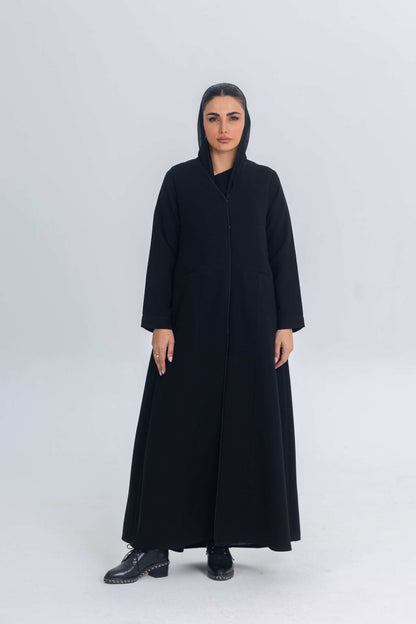 Woman wearing black crepe abaya with subtle satin detailing and hood, standing against plain background