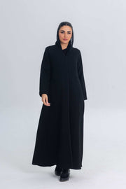 Woman wearing black crepe abaya with subtle satin detailing and hood, standing against plain background