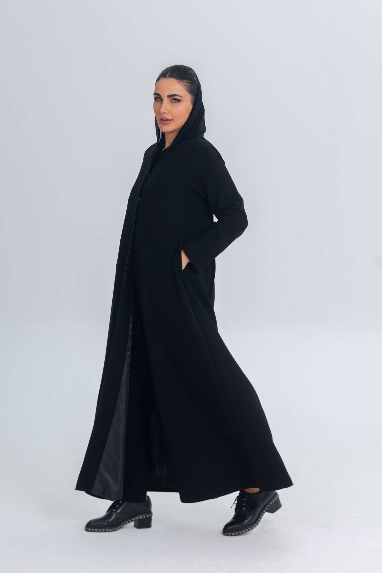Woman wearing Noir Elan black crepe abaya with satin detailing and hood, styled with black studded boots.