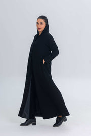 Woman wearing Noir Elan black crepe abaya with satin detailing and hood, styled with black studded boots.