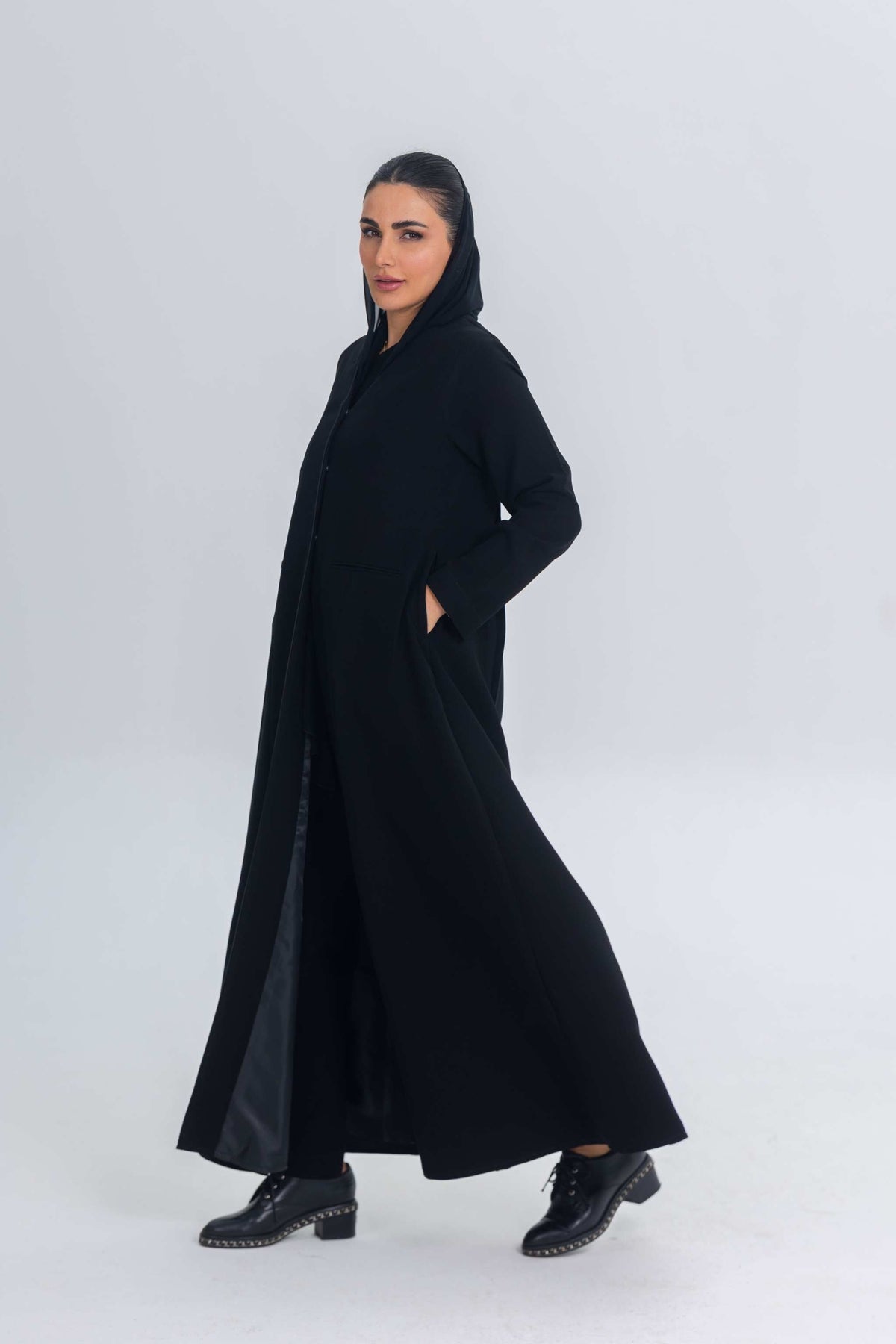 Woman wearing Noir Elan black crepe abaya with satin detailing and hood, styled with black studded boots.