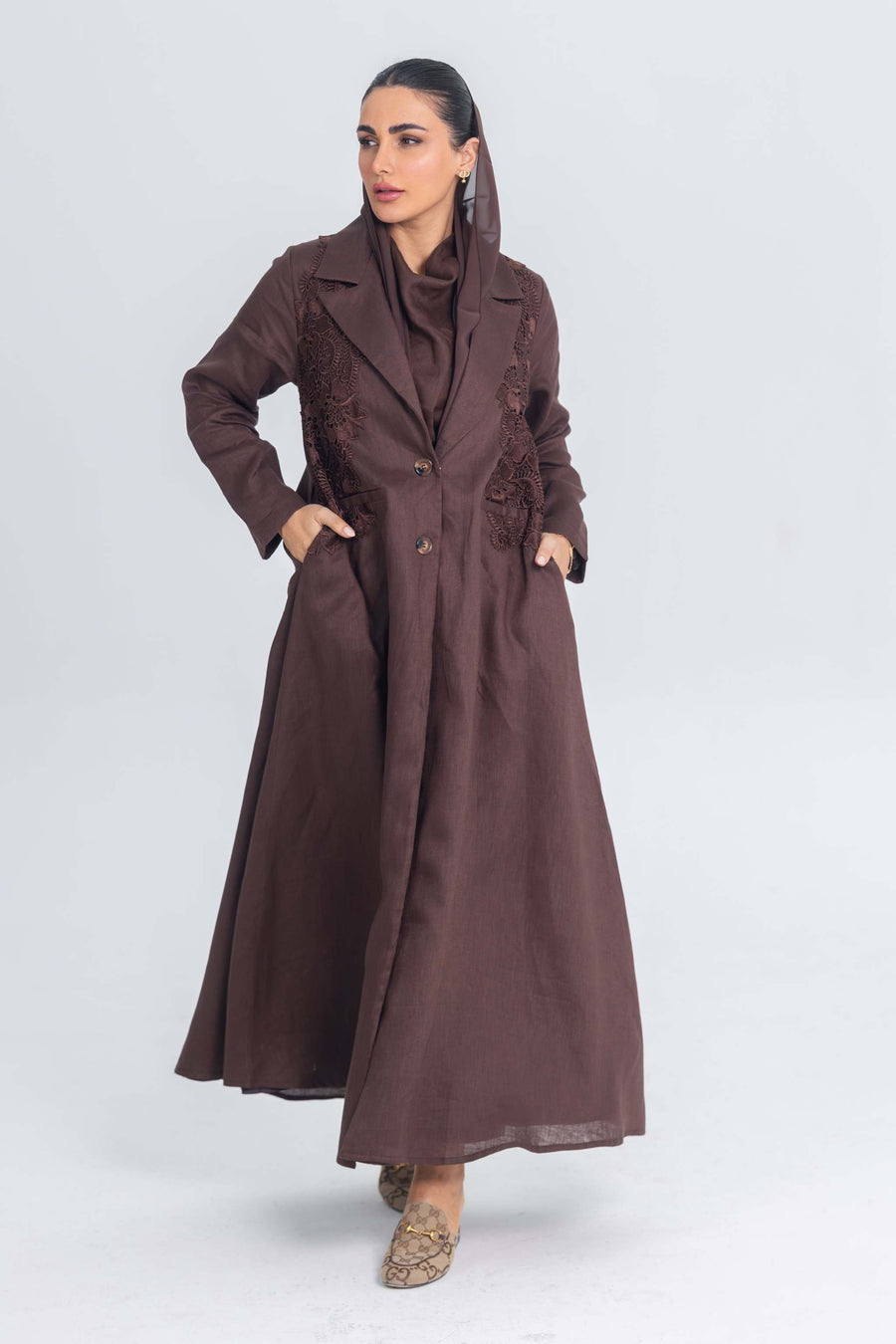 Woman wearing brown linen abaya with blazer collar and floral lace embroidery patches, standing against light background
